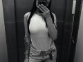 Black-and-white selfie of Olieri with long dark hair, sleeveless white top, showing small tits, high-waisted jeans, tattoo on right arm, holding phone in front.