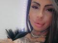 DenyChantal with tattoos, wearing a black feathered garment, has full lips, a nose ring, and a gem necklace, smiling at the camera with dark eyebrows and long brown hair.