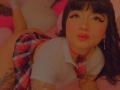 AnnieGrey with dark hair, eyeliner, and red lipstick, wearing a plaid skirt, white shirt, and red choker, lying on a pink bed, showing tattoos on her arms.