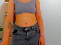 Steisii has a flat stomach, small boobs, and a tattoo on her right arm. She's wearing a gray sports bra and high-waisted, dark blue jeans.