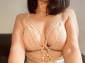Close-up photo of LorenHolmes, wearing a sheer, beige lace bralette that accentuates her large breasts. Her dark bob haircut partially covers her face. The bralette has thin straps and a central clasp.