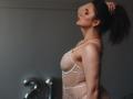 Dreia, com a cabeça inclinada para trás, vestindo um vestido branco de renda transparente, tem a mão no rabo de cavalo castanho, exibindo seus pequenos seios e delicadas joias..