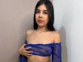 NaomyRosse with long black hair, red lipstick, and blue sheer top, covering her small tits with her hand. She's standing against a white and beige wall.