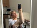 ArnettaNazario taking a selfie, mouth open, tongue slightly out, blonde hair in a clip, wearing a beige cardigan, white shirt, and black tights, holding a black phone.