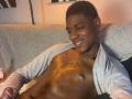 DrakeWit, shirtless, with a shiny, muscular, dark brown chest, wearing a chain necklace and a gray open hoodie, smiling at the camera. Short, curly hair, relaxed, on a couch.