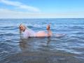 FallenAngelia, lying in shallow water, face down, wearing a wet, see-through shirt, exposing her ass and bare back, with blonde hair, legs bent, feet in air.