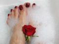 VanasMoka's feet in a bubble bath, with red-painted toenails and a red rose resting between her toes. Her skin is pale, and the rose is fresh.