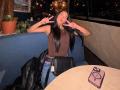 JennTylor with long black hair, brown sleeveless top, blue jeans, black purse, making double peace signs, mouth open, black jacket on table, phone on table, night outside.