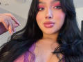 LilyConorr with long black hair, pink eyeshadow, glossy pink lips, and purple lace top, showing off her tits and shiny nails, smiling at the camera.