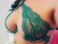 FrenchShanna's tits in a green lace bra, showing side profile with visible tattoo below her right breast, dark curly hair, and tan skin.