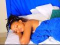 Sashie with dark skin, closed eyes, and braided hair sleeps on a bed, covered in a blue towel, with white and green pillows. Sashie's ass is partially visible.