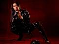 AfroditaDoir vêtue d'une combinaison en latex noir, accroupie, suçant son doigt à lèvres rouges, bottes brillantes, talons aiguilles, fouet sur le sol rouge, fond rouge, longs cheveux noirs, séduisante, sexy..