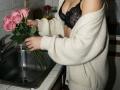 Destinis with pink roses, washing them under a kitchen sink, wearing a black lace bra and a loose, off-shoulder white sweater. Boobs partially visible, hand holding roses.