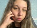 Sanella with puffy pink lips, green eyes, and brown wavy hair, holding a phone to her ear, looking bored. She's got a slight acne mark on her forehead.