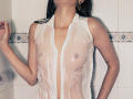 JennaDerose with wet white shirt, showing her tits and nipples, tattoo on left arm, dark hair, gold earrings, standing against white tiled wall.