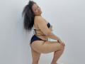 KloheEvans has curly black hair, tan skin, and is squatting in black bikini, showing her big ass and small tits, smiling at the camera.