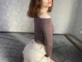CicelyDence with shoulder-length brown hair, pink lips, wearing a tight, off-shoulder purple top and white, frayed denim shorts, kneeling on a gray bed. Boobs, ass, top, shorts.
