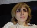 EmelyDilsaver has blonde shaggy hair, light skin, and a slightly pouty face with a small mole on her cheek, wearing a white shirt, looking to the side.