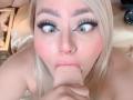 Foto em close-up de RoxannEmilss, uma mulher loira com olhos verdes, delineador marcante e um piercing no nariz, chupando um grande... pênis circuncidado, ereto, com a mão direita dela envolvendo-o..