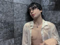 RubyMote with wet black hair, wearing a sheer white shirt, exposing small tits and a silver necklace, leaning against dark gray tiled wall, looking up with a slight smile.