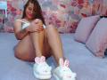 BelletinaXx with brown hair, red lipstick, white bra, no panties, slippers with bunny ears, sitting on bed, hugging knees, showing her ass and tattoos, red nails.