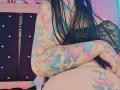Kkole with long black hair, wearing black lace, has colorful tattoos on arms and thigh, and is sitting with crossed arms, showing a cherry tattoo on her thigh.