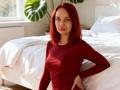 BrendaBeautys with red hair, wearing a red long-sleeve shirt, sitting by a white bed, hands on her hips, smiling softly, medium tits, light skin, bedroom.