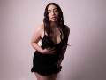 AnnFisher with long wavy brown hair, wearing a black dress with a low neckline, showing her tits, holding her dress, standing against a plain background, looking at camera.