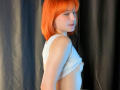 MasakoParkey with bright orange bob has pale skin, wearing a white tank top, exposing her small tits, looking back over her shoulder, black curtain background.