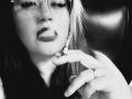 BeccaVanBoa with glasses, dark lipstick, and a ring, holding a cigarette to her mouth, her finger pointing slightly, black and white, she's got a pouty face.