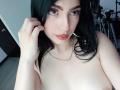 IsabellaaRossi with black hair, green eyes, pink eyeshadow, red lipstick, and a silver necklace, topless, showing her small tits, hand touching her hair, in a selfie.