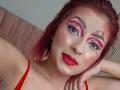 LizBonnet has red makeup, pink eyeshadow, red lipstick, and red eyebrows with a small pink heart tattoo above her right eyebrow. She's wearing a red top, looking at the camera.