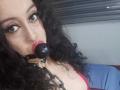 Kendra Simons, met krullend zwart haar, bruine ogen en rode lippenstift, draagt een zwarte ball gag en ketting, kijkt omhoog met een licht geopende mond, waarbij een beetje van haar borsten zichtbaar zijn..