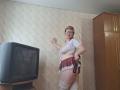 SabrinaShine with glasses, white crop top, red plaid mini-skirt, white lace thigh-highs, and black sex toy, standing, hand on wall, TV on left, wooden cabinet on right.
