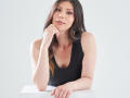 KarlaMazuera with long brown hair, wearing a black sleeveless top, leaning on a white surface, hand under chin, looking at camera with subtle smile, light makeup, soft expression.