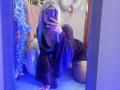 MaddieLesYoux with blue hair, wearing a black lace dress, on all fours, taking a mirror selfie, showing her tits and tattoo on her thigh, in a blue-lit room.