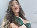 TaniaSandersX with long wavy brown hair, green lace bra, and pouty red lips, looking down at the camera, showing her tits. Soft light, slightly blurry background.