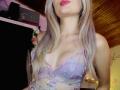 EliseMonet, avec ses cheveux blonds, son rouge à lèvres rouge et son soutien-gorge en dentelle florale violette, montrant ses seins, dans une pièce faiblement éclairée avec un plafond en bois..