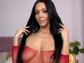Nadeen with long black hair, wearing a red mesh top that reveals her big tits, smiling, holding her hair with both hands. She has a gold necklace and ring.