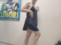 JoannaSia wearing a black, sleeveless, side-strap dress, exposing her ass and upper thighs, with a hand on the wall, in a room with a colorful poster.