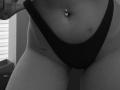 SugarrX's black thong, showing a belly button piercing with two small jewels, and a tattoo just above their thigh. Black-and-white photo, focusing on their lower torso.