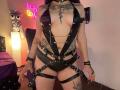LilithDark69 with purple hair, black leather strappy outfit, showing tits, tattoos, and black straps, standing in a bedroom with a neon sign and a lit lamp.