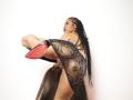 LuciaVargasX with long black braids, lifting one leg in a sheer black lace dress, showing her ass and red-soled high heel, leaning on a brown chair.