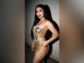 MiilaWx with long black hair, wearing a gold sequined strapless dress with chain details, standing in a shadowed area, showing her ass and a confident smile.