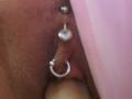 FrenchShanna's pussy with three silver piercings: a ball, a ring, and a small diamond. The skin is brown and slightly textured. A pink surface is in the background.