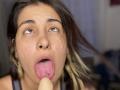 NyxVale's face with brown eyes, nose ring, and tongue out, has a tongue piercing and is sucking a large, flesh-colored dildo. Her dirty blonde hair is slightly messy.