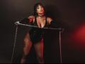MadisonHetfield with tattoos, black corset, fishnet stockings, and gloves, holding chains, leaning forward, red and black lighting, confident expression, visible boobs, and chain around neck.