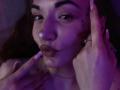 LelaTraverso's face with purple lighting, pouty lips, and hands touching her head. She has a nose piercing, dark eyebrows, and a small tattoo near her cheek.