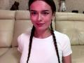 DrusillaMaiava with braided brown hair, pinkish skin, white t-shirt, small nose piercing, slight smile, sitting on beige leather couch, small brown cat statue in background.