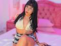 GraceClarke with long black hair, glasses, and a yellow crop top, showing her small tits, wearing a plaid skirt, sitting on a bed, pink lighting, looking at the camera.